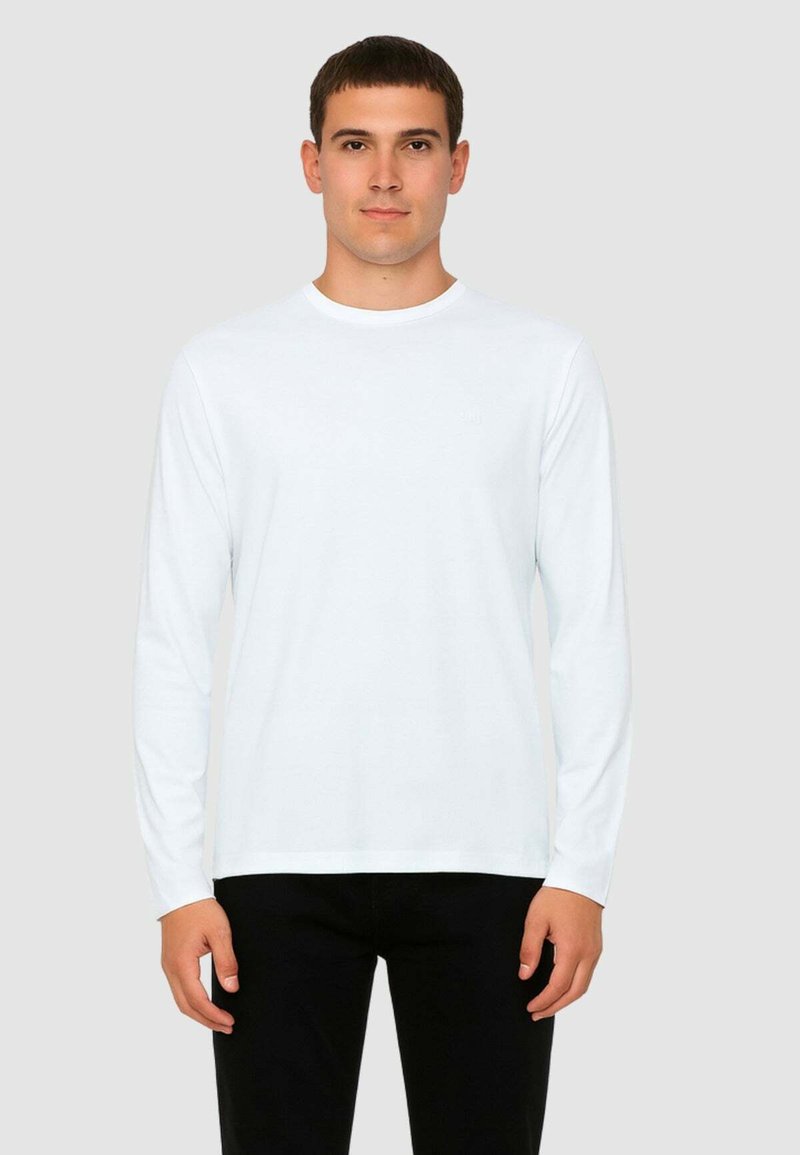 Camicia bianca a maniche lunghe in un tessuto liscio con un classico scollo rotondo e un logo del brand discreto sul petto, indossata con pantaloni neri.