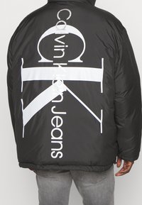 Black puffy jacket featuring large white branding on the back, with "Calvin Klein Jeans" text and geometric patterns. Smooth texture.