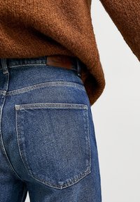 Jeans in denim blu scuro con superficie texturizzata, dotati di una patch in pelle marrone in vita e due tasche posteriori.