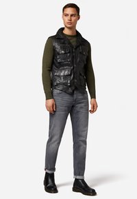 Schwarze Lederweste mit mehreren Taschen, mit einer leicht strukturierten Oberfläche. Getragen über einem dunkelgrünen Langarmshirt und grauen Jeans.
