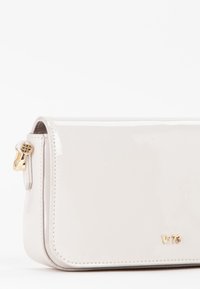 Bolso beige brillante con forma rectangular, herrajes dorados y bordes cosidos, que presenta un logo en la parte frontal.