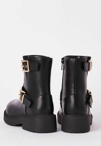 Botas de tobillo de cuero negro con hebillas en tono dorado, punteras redondeadas y suelas de goma bajas. Incluyen un cierre de cremallera y un acabado texturizado.