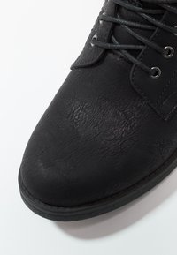 Zapato negro con cordones, confeccionado con un acabado de cuero sintético texturizado, punta redonda y suela de goma. Presenta cordones grises y ojales metálicos.