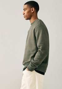 Olivgrön sweatshirt med ribbad rundhals och muddar, tillverkad av textil med struktur, ihop med ljusbeiga byxor. Modellen står på sidan.