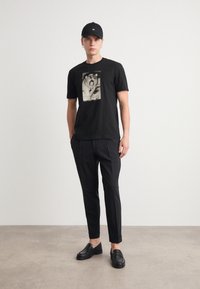 Tricou negru cu un imprimeu grafic, combinat cu pantaloni negri tăiați și pantofi slip-on negri. Modelul poartă o șapcă neagră.