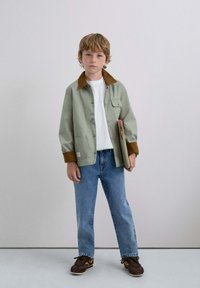 Jachetă verde cu guler maroniu, tricou alb, blugi albaștri și adidași maronii. Băiat ținând un caiet, stând pe un fundal simplu.
