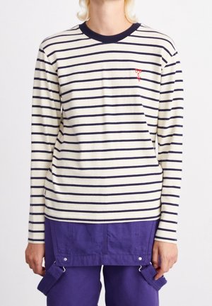 Personne portant une chemise blanche à manches longues avec des rayures horizontales bleu marine et un petit logo rouge brodé, associée à un pantalon violet et une ceinture utilitaire.