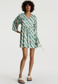 Kurzes Wickelkleid in Creme mit grünem Batikmuster. Merkmale sind eine geraffte Taille, ein schwingender Rock und eine Bindeschnur an der Seite.