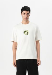 T-shirt en coton blanc avec un col rond et des manches courtes. Présente un graphique de soleil vert et noir avec un visage souriant sur la poitrine.