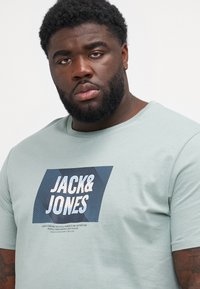 Ανοιχτό πράσινο βαμβακερό μπλουζάκι κοντό μανίκι, με μπλε και λευκό γραφικό σχέδιο που περιλαμβάνει το κείμενο "JACK&JONES" και λωρίδες.