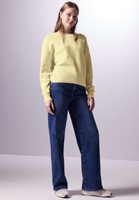 Lichtgele geribbelde gebreide trui met lange mouwen, gecombineerd met wijde donkere blauwe spijkerbroek en grijze sneakers, tegen een neutrale achtergrond.