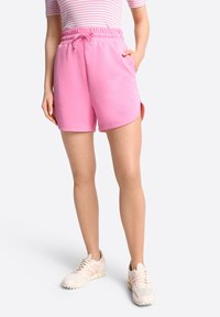 Růžové šortky z bavlněné směsi s elastickým pasem a šňůrkou, s bočními kapsami a zaobleným lemem. V páru s béžovými teniskami.
