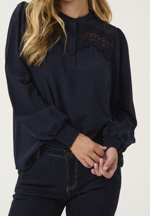 Vrouw draagt een donkerblauw blouse met kanten details en stoffen knopen, gecombineerd met donkere denim jeans, handen zachtjes in elkaar gevouwen vooraan.