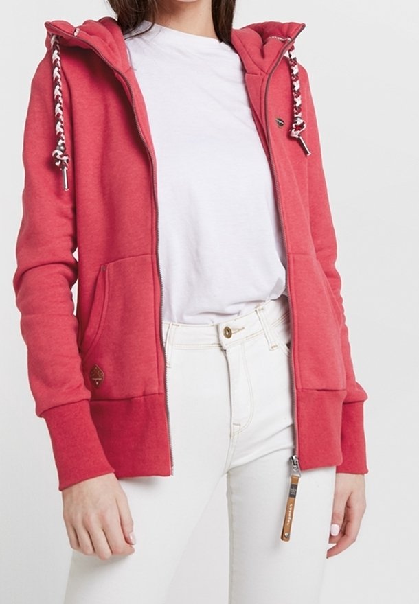 Rode zip-up hoodie met een trekkoord capuchon, twee voorkruiszakken en witte accenten. Draagt over een wit shirt, gecombineerd met lichtgekleurde broek.