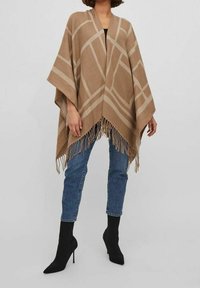 Beżowy i brązowy ponczo w wzory z frędzlami, z geometrycznym wzorem. Nosi się je na czarnej bluzce, w zestawieniu z ciemnymi dżinsami i botkami za kostkę.
