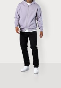 Felpa con zip viola chiaro con logo, abbinata a jeans neri strappati e sneakers bianche, su uno sfondo neutro.