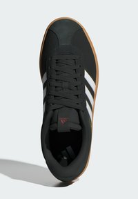 Crna atletska cipela s bijelim i zlatnim prugama, gumeni potplat, teksturirana gornja tkanina i crne vezice. Sadrži logo na jeziku.