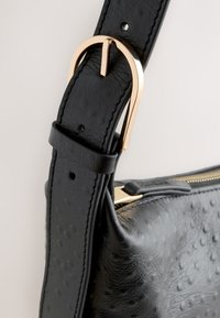 Black textured leather bag strap with gold buckle and adjustable holes, attached to a matching textured leather bag with a zipper.