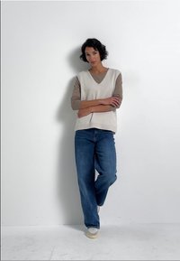 Ljus beige ärmlös topp över en tan långärmad tröja, kombinerat med lösa blå jeans och vita sneakers; enkel, avslappnad passform.