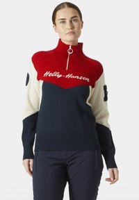 Sudadera de punto con cierre en rojo, blanco y azul marino con un diseño en forma de chevrón. Cuenta con un logo y puños y dobladillo acanalados. El material es suave y texturizado.