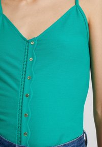 Bodysuit sin mangas de color verde azulado con un escote en V, que presenta una fila decorativa de botones dorados y un borde ondulado a lo largo de la abertura frontal.