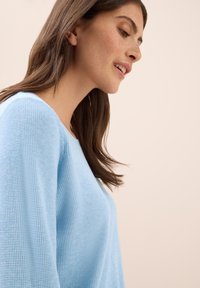 Maglione azzurro chiaro con un tessuto a maglia testurizzato. Ha maniche ampie e un collo rotondo. Il modello è leggermente girato di lato.