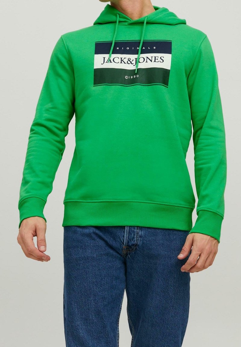 Muž v jasně zelené mikině s logem "JACK & JONES" a tmavě modrých džínách, stojící proti jednoduchému světlému pozadí.