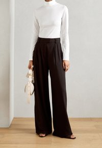 Valkoinen poolopaita yhdistettynä korkeavyötäröisiin tummiin avariin housuihin. Korostettuna vaalealla käsilaukulla ja nyörisaappailla puulattialla.