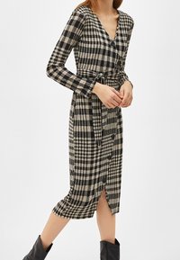 Svart och beige rutigt klänning med långa ärmar, V-ringning, bälte i midjan och en sidoklyv. Tillverkad av vävt material med knappdetaljer.