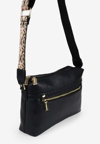 Bolso cruzado de cuero negro con cremalleras doradas, superficie texturizada y una correa con un patrón en rosa y negro. Presenta dos compartimentos con cremallera.