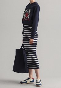 Person som bär en marinblå sweatshirt med ett emblem, en marinblå och vitrandig midi-skjol, sneakers och håller en stor marinblå tygväska, stående i profil.