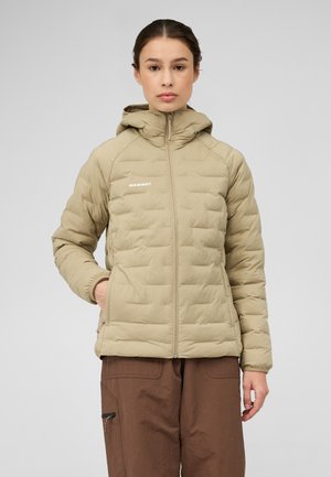 Vrouw die een beige gewatteerde Mammut-jas met capuchon draagt en een bruine broek staat met één hand in haar zak tegen een grijze achtergrond.