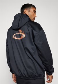 Giacca nera con cappuccio realizzata in tessuto liscio. Presenta un design di fiamme colorate sul retro e polsini elasticizzati all'orlo e alle maniche.