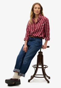 Chemise en flanelle à carreaux rouges et blancs avec les manches retroussées, associée à un jean bleu clair ample et des chaussures foncées, assise sur un tabouret en bois.
