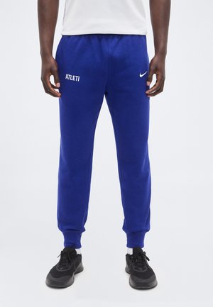 Homme portant un jogging bleu avec les logos "ATLETI" et Nike, associé à des baskets noires et des chaussettes blanches, debout devant un fond uni.