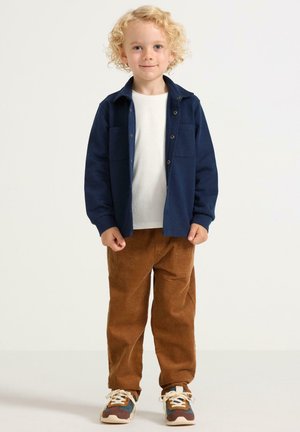 Bambino con i capelli biondi ricci, che indossa una giacca blu navy, una camicia bianca, pantaloni di velluto a coste marroni e scarpe da ginnastica multicolori, in piedi su uno sfondo bianco.