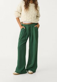 Persoon in een crèmekleurige kabelgebreide trui, losse groene broek met trekkoord en beige sneakers, staande met handen in de zakken.