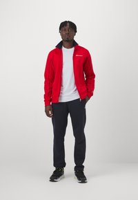 Chaqueta deportiva roja con cremallera y cuello oscuro, camiseta blanca debajo, pantalones de chándal azul marino y zapatillas negras con detalles verdes.