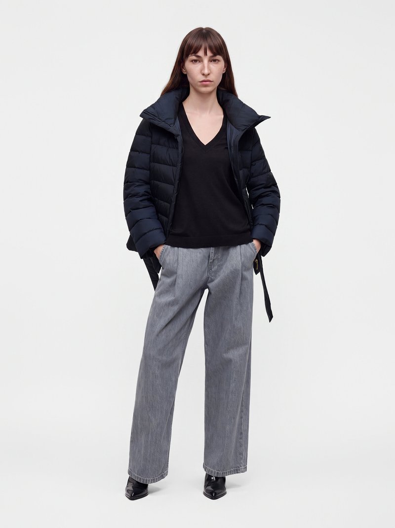 Femme portant une doudoune noire, un pull noir à col en V, un pantalon large gris et des chaussures noires pointues, debout les mains dans les poches.