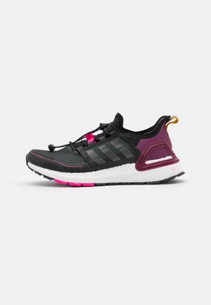Chaussure de sport noire et violette avec semelle blanche texturée, détails roses, lacets noirs et languette jaune sur fond uni.