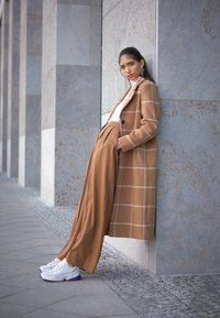 Brun rutigt kappor över en vit polotröja, ihop med beige vida byxor och vita sneakers med lila detaljer, mot en strukturerad vägg.