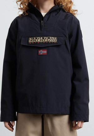 Personne portant une veste-pull Napapijri bleu marine avec col zippé et écusson du drapeau norvégien, associée à un pantalon beige clair.