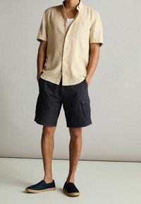 Camisa de lino corta de manga en beige claro, pantalones cortos de carga azul marino con bolsillos laterales y zapatos oscuros sin cordones con suela de yute trenzada.