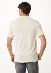 Jasnobeżowa koszulka polo z krótkim rękawem, z klasycznym kołnierzykiem, prążkowanymi mankietami i gładką fakturą, włożona w szare dżinsy.