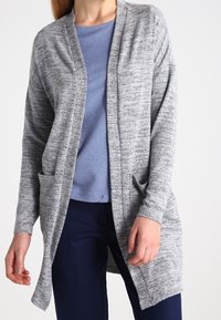 Cardigan grigio mélange con colletto a scialle, maniche lunghe, tasche frontali e apertura frontale. Indossato sopra una maglietta blu e pantaloni scuri.