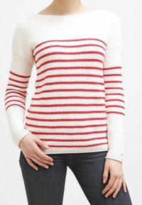 Weißer Strickpullover mit roten horizontalen Streifen, weitem Ausschnitt, langen Ärmeln und gerippten Bündchen. Getragen mit dunklen Jeans.