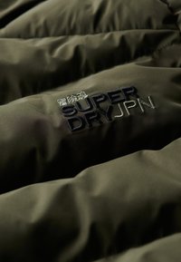 Giacca puffer verde oliva in tessuto con una superficie liscia e leggermente lucida. Presenta il logo ricamato "SUPERDRYJPN" in blu navy e verde chiaro.