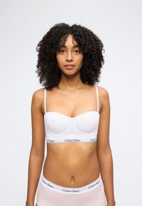 Γυναίκα με μπούκλες που φοράει λευκό σουτιέν Calvin Klein και ταιριαστό εσώρουχο, μπροστά σε απλό γκρι φόντο.