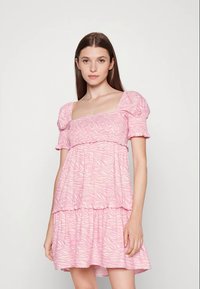 Jovem mulher com cabelo castanho longo, vestindo um vestido curto de mangas curtas, em camadas, com um padrão ondulado subtil, apoiada contra um fundo branco.