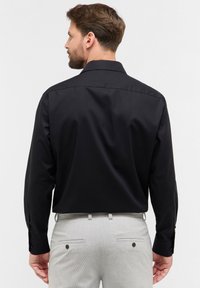 Camicia nera a maniche lunghe con collo classico, tessuto liscio e polsini con bottoni; indossata con pantaloni grigio chiaro dotati di tasche posteriori.